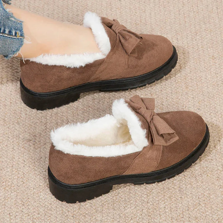 Brown suede slip-on shoes with white faux fur lining and decorative bow on a beige carpet
