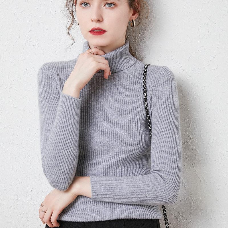Young woman wearing a gray ribbed turtleneck sweater posing against a textured white wall