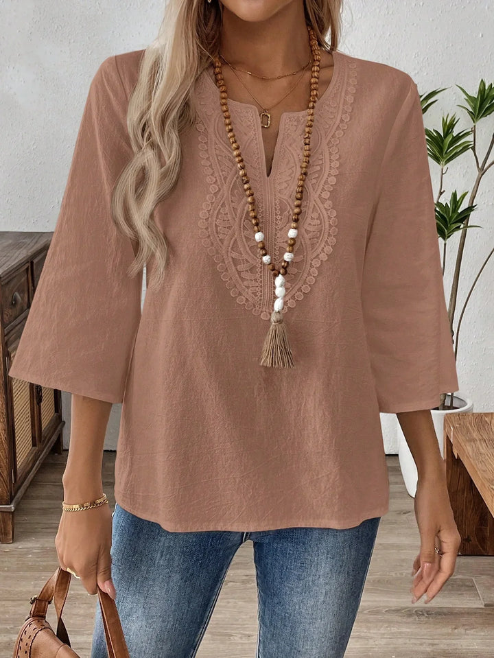 Woman wearing a dusty pink embroidered blouse with 3/4 sleeves and blue jeans, accessorized with a wooden bead necklace