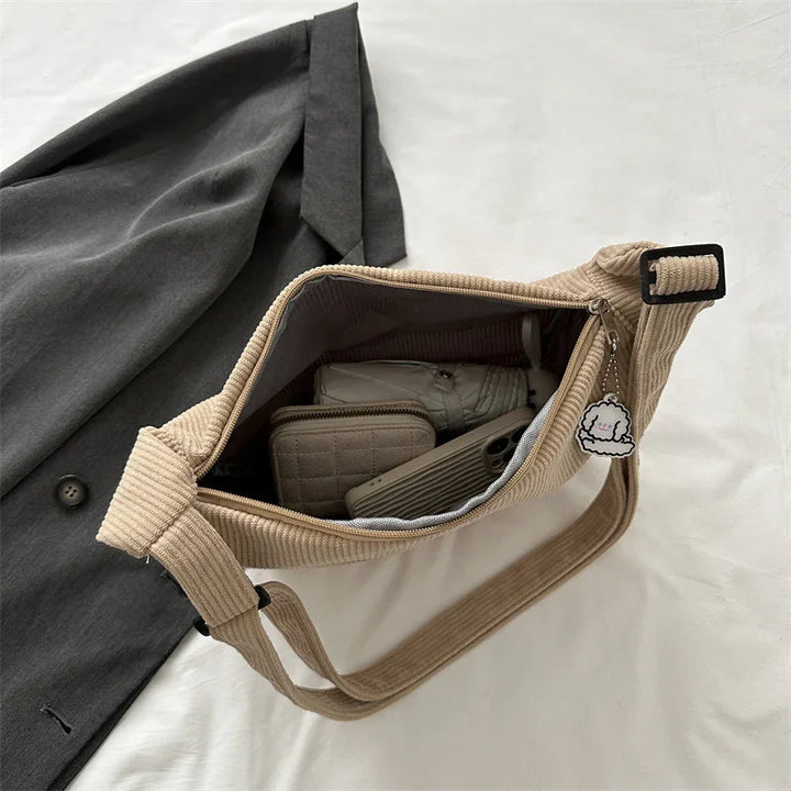 Beige corduroy crossbody bag with zipper, keychain, containing wallet, shoes, and phone on white surface next to dark gray coat