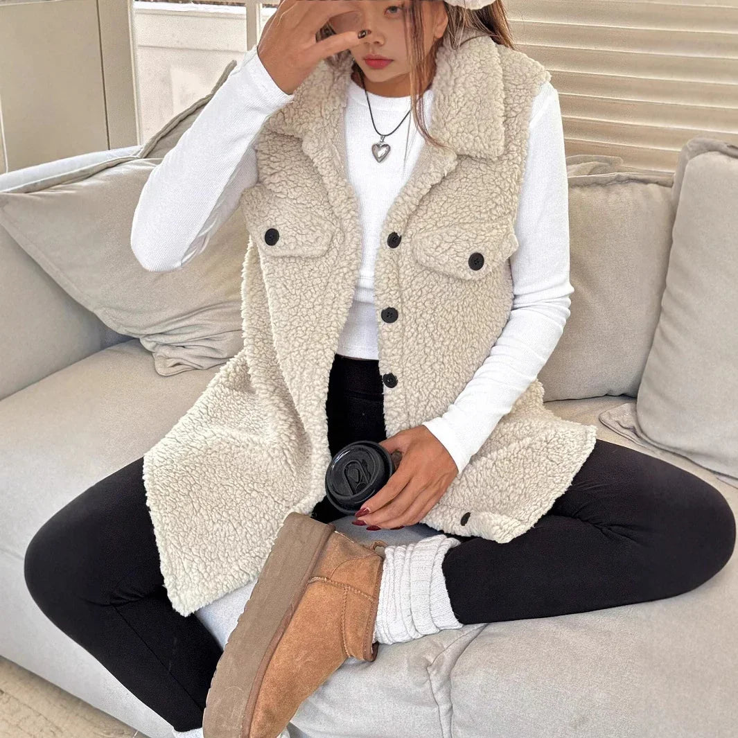 Woman seated on beige sofa wearing cream sherpa vest, white long sleeve shirt, black leggings, and tan ankle boots