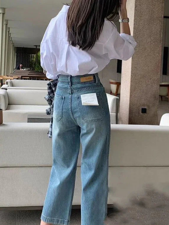 Woman wearing light blue denim jeans with a white shirt, standing indoors near beige sofas