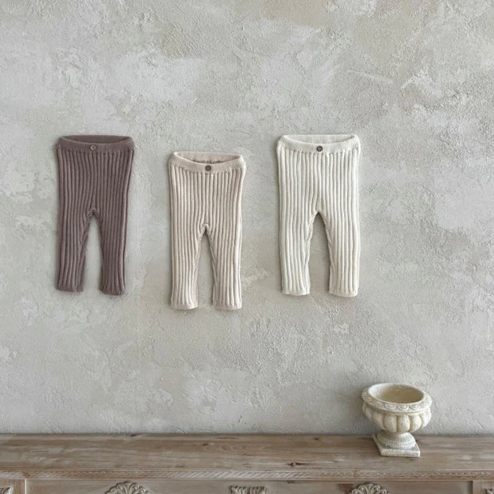 Three pairs of ribbed knit baby leggings in brown, beige, and white hanging on a textured wall above a wooden table with a decorative stone planter