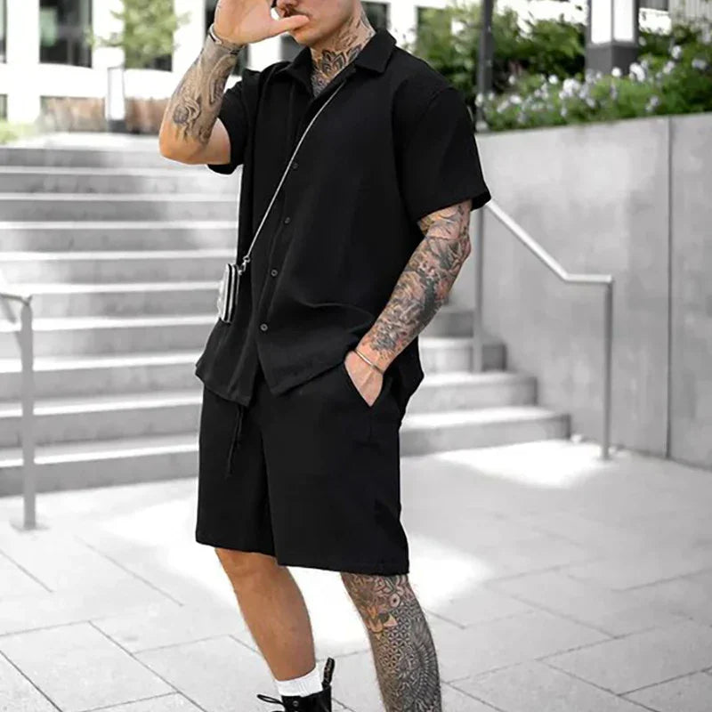 Man wearing black short-sleeve button-up shirt and matching shorts with visible tattoos outdoors