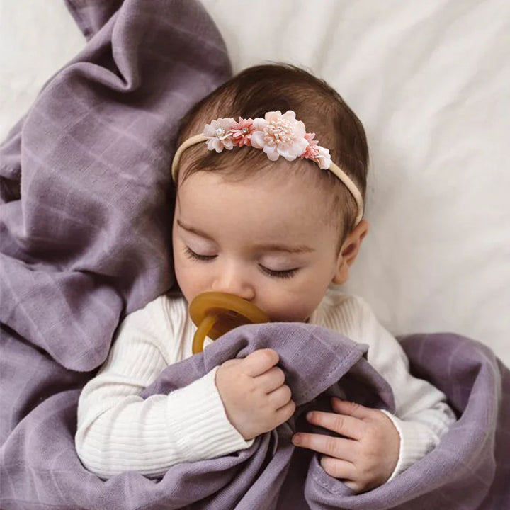 Sleeping baby with floral headband and pacifier wrapped in soft purple blanket on white bed