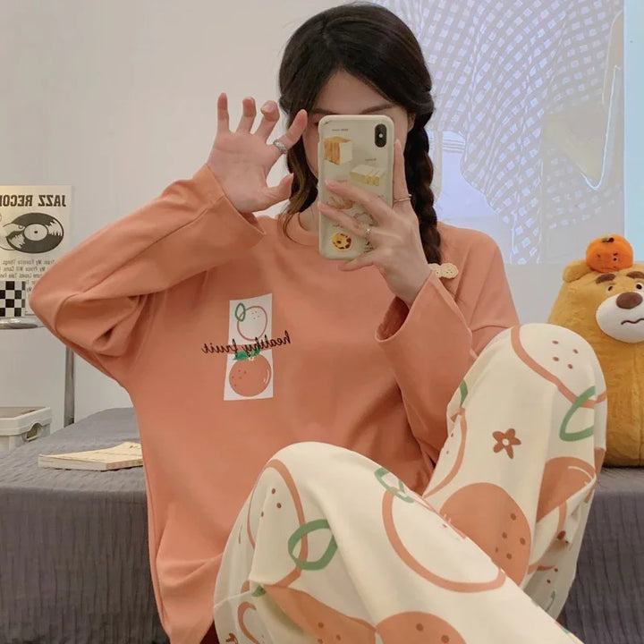 Woman in peach long-sleeve shirt with fruit graphics and patterned pants taking mirror selfie in cozy bedroom