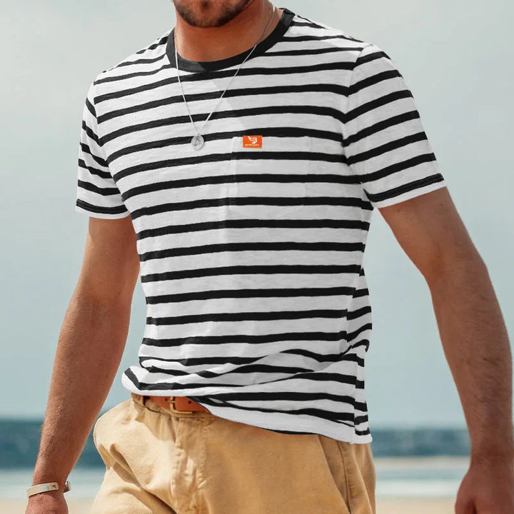 Man wearing black and white striped t-shirt with orange logo, beige pants, and silver necklace at the beach