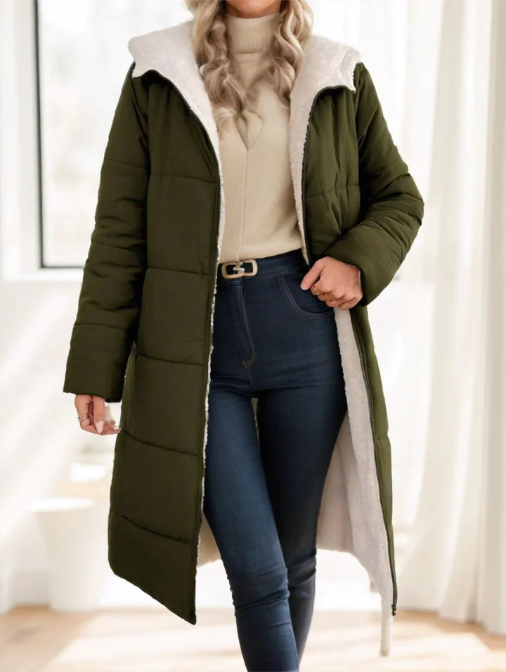 Woman wearing olive green long puffer coat with white fleece lining and dark blue jeans indoors