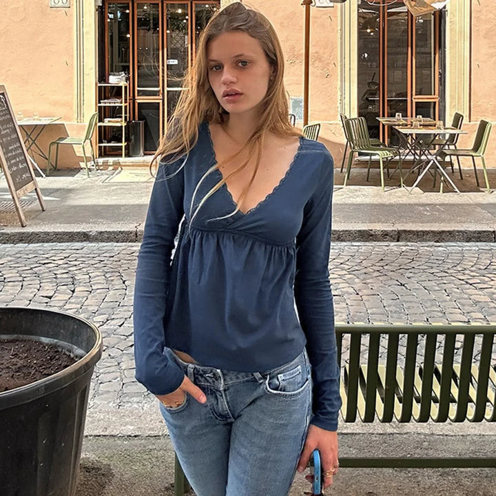 Young woman in blue long-sleeve top and jeans standing near green bench outside cafe on cobblestone street