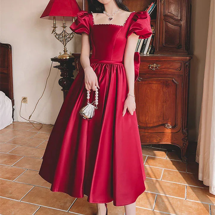 Woman in elegant red satin puff sleeve midi dress holding silver pearl handle handbag indoors