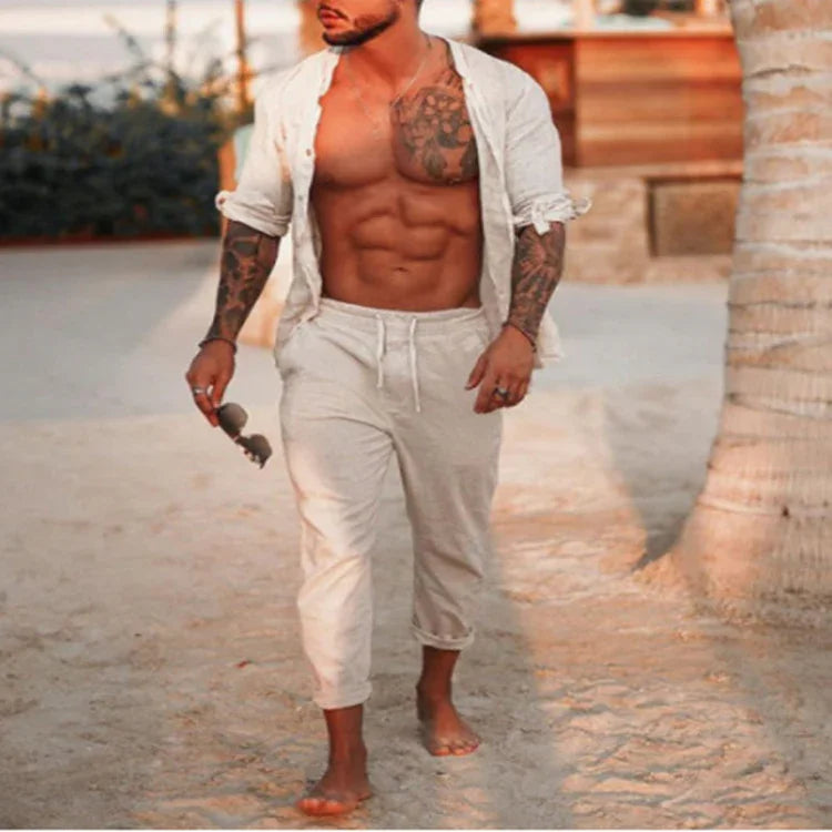 shirtless muscular man with tattoos walking barefoot on sandy beach wearing beige pants and open shirt