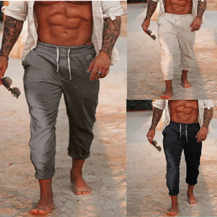 Fit man with tattoos wearing rolled-up drawstring pants in gray, beige, and black walking barefoot on sandy beach