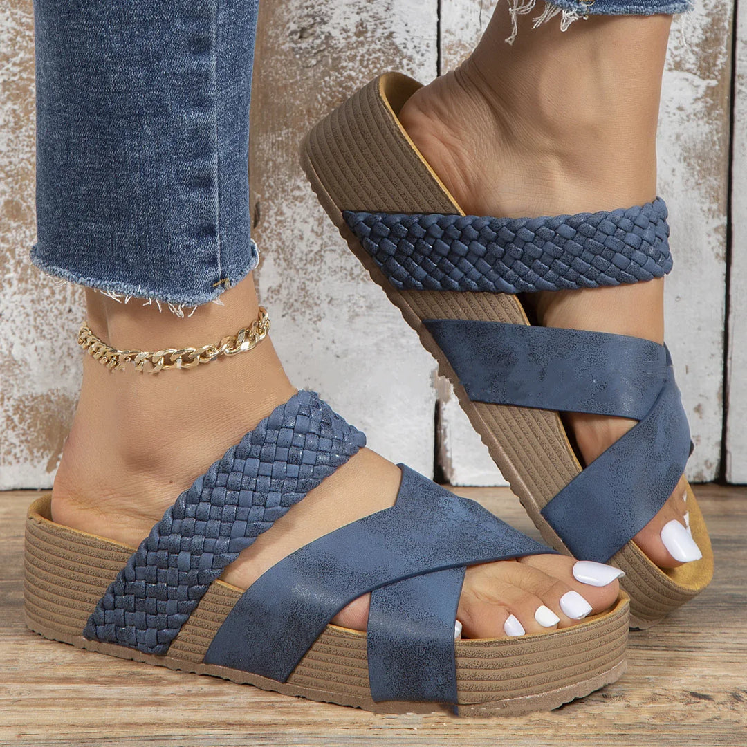 Close-up of feet wearing blue wedge sandals with braided and smooth straps on wooden floor