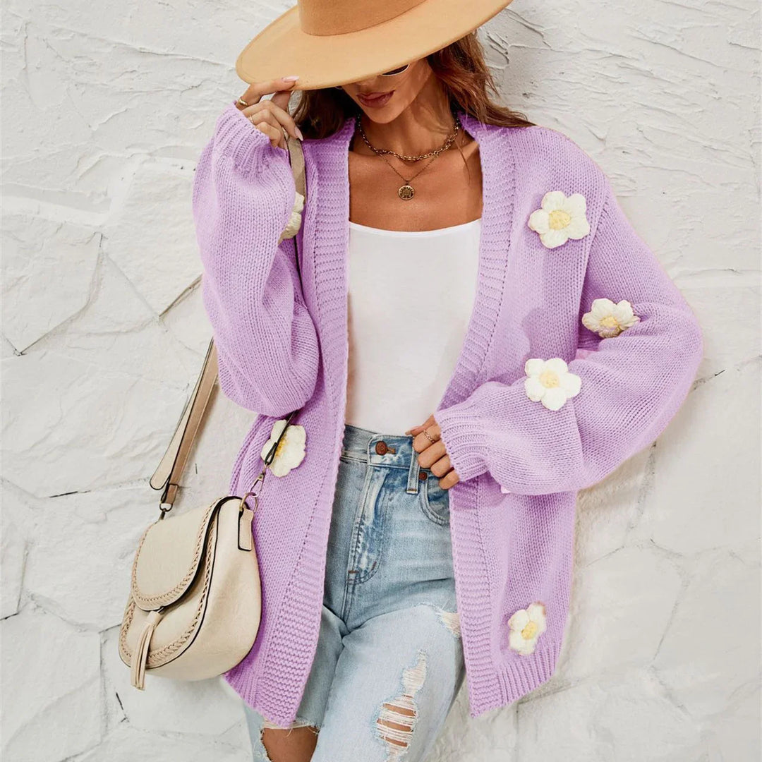Woman wearing lavender knit cardigan with white flower accents, white tank top, ripped jeans, beige hat, and cream handbag