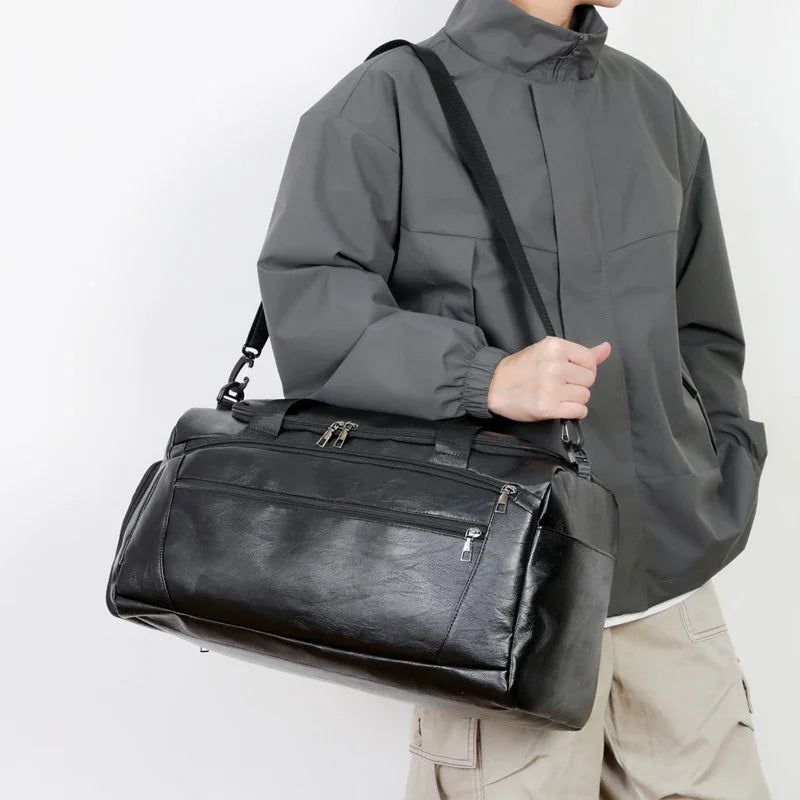 Person wearing gray jacket and beige pants holding black leather duffel bag with shoulder strap
