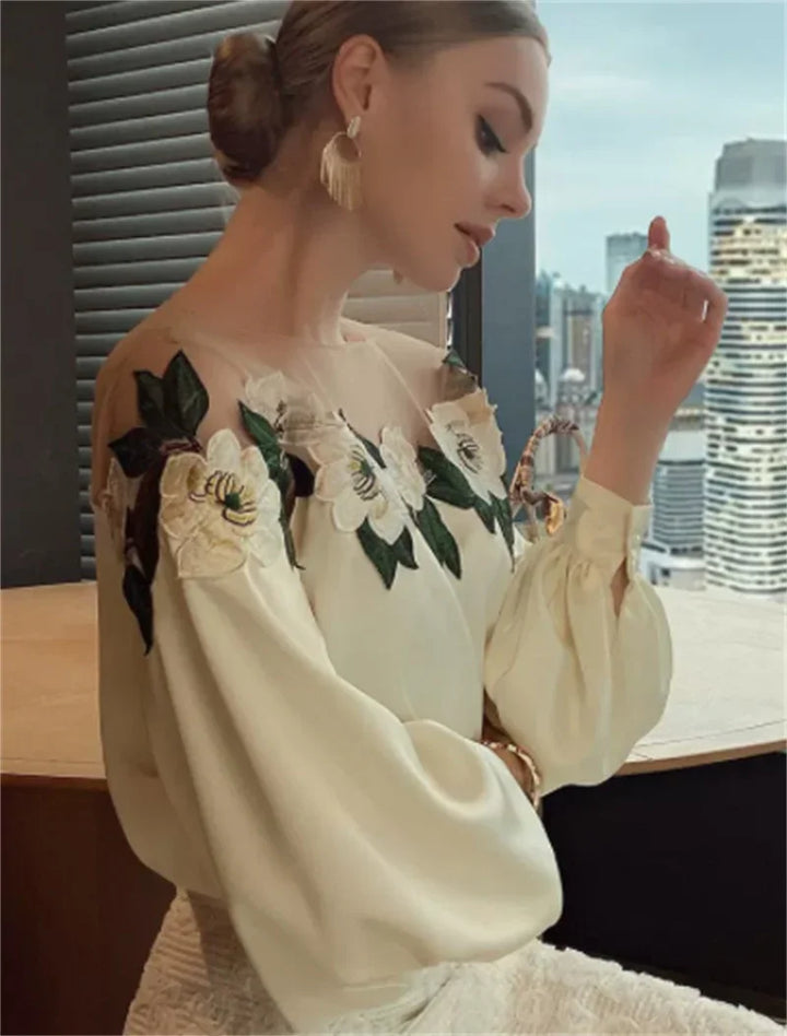 Elegant woman wearing cream blouse with floral embroidery sitting by window overlooking cityscape