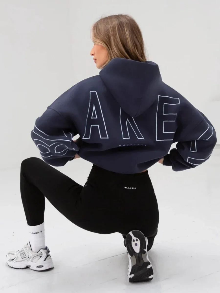 Woman wearing navy cropped hoodie and black leggings, white sneakers, posing in studio