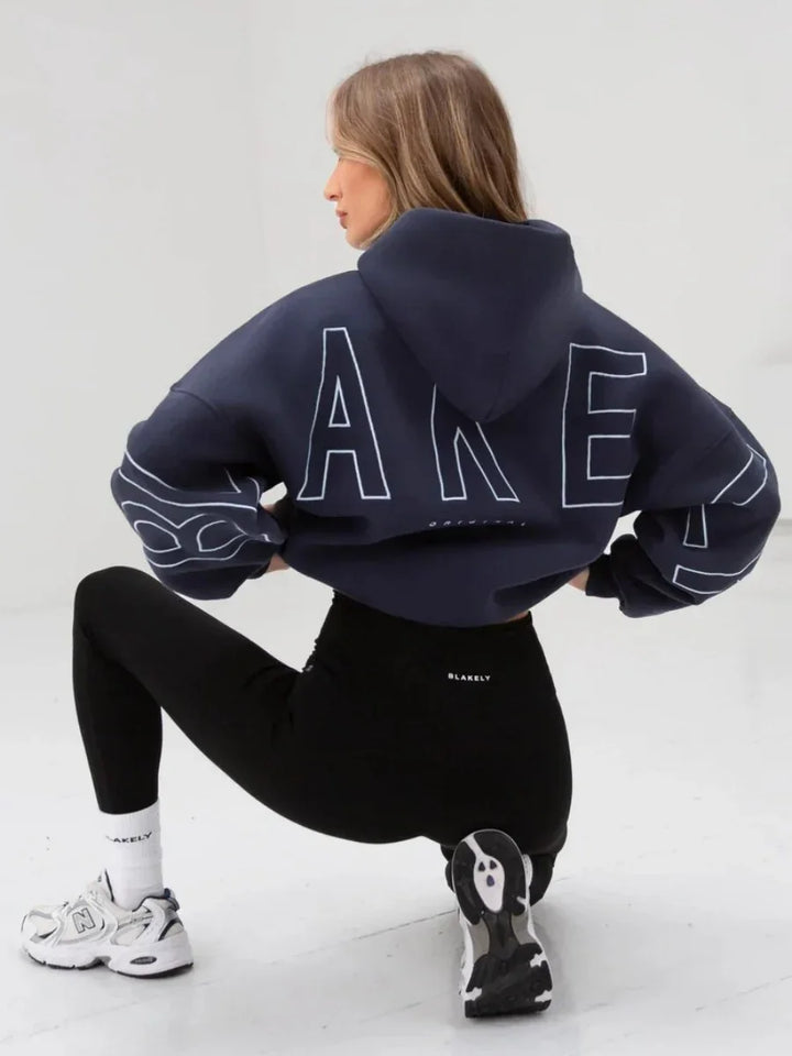 Woman wearing navy cropped hoodie and black leggings, white sneakers, posing in studio