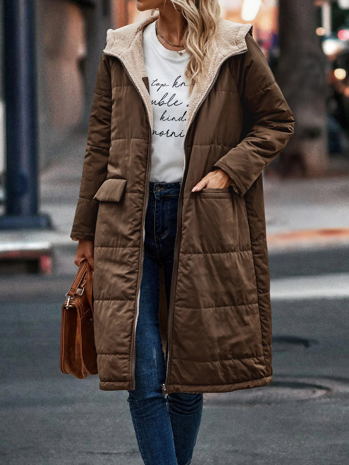 Woman wearing long brown padded coat with sherpa lining, casual jeans, white graphic tee, and carrying brown handbag on city street