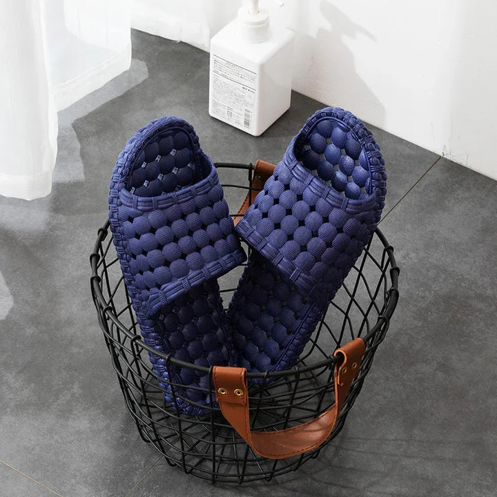 Pair of navy blue textured slippers in black wire basket with brown leather handles on gray floor