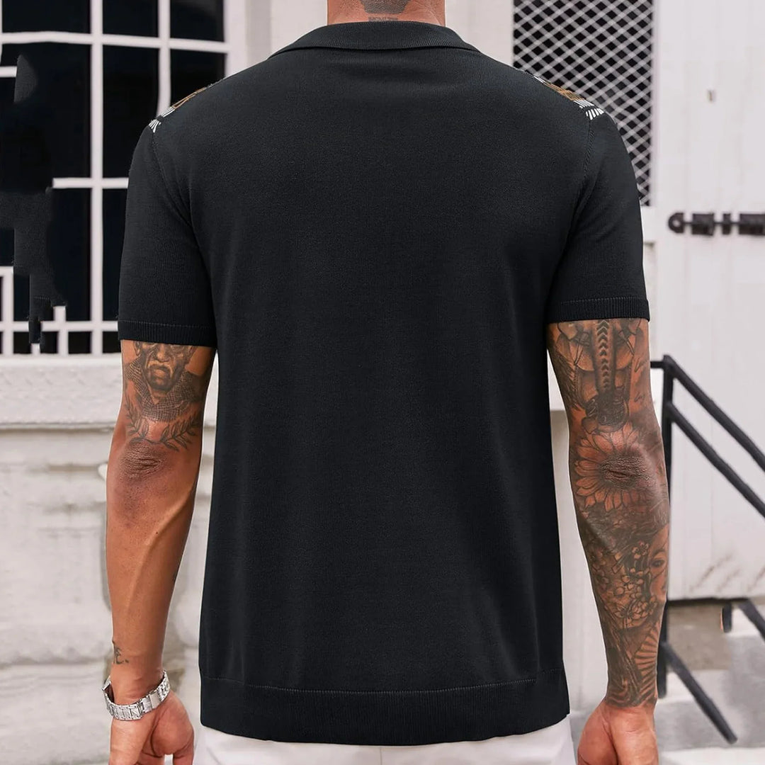 Man wearing black short-sleeve knit shirt showing tattooed arms against urban backdrop
