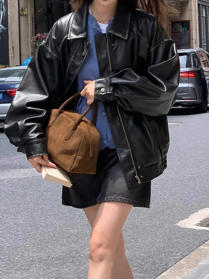 Woman wearing black oversized leather jacket and black skirt, holding brown suede handbag and book on city street