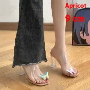 Pair of feet wearing transparent high-heeled sandals with colorful butterfly decorations on wooden floor