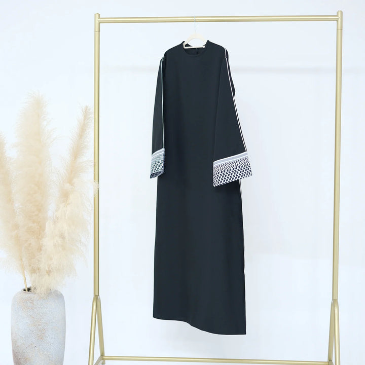 Black abaya with white patterned cuffs hanging on gold clothing rack beside pampas grass vase
