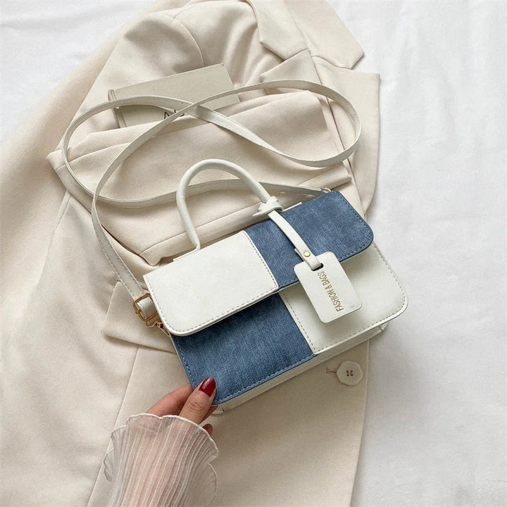 Hand holding a stylish blue and white colorblock handbag with top handle and shoulder strap on beige blazer