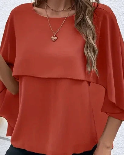 Woman wearing a rust orange layered blouse with flutter sleeves and heart pendant necklace
