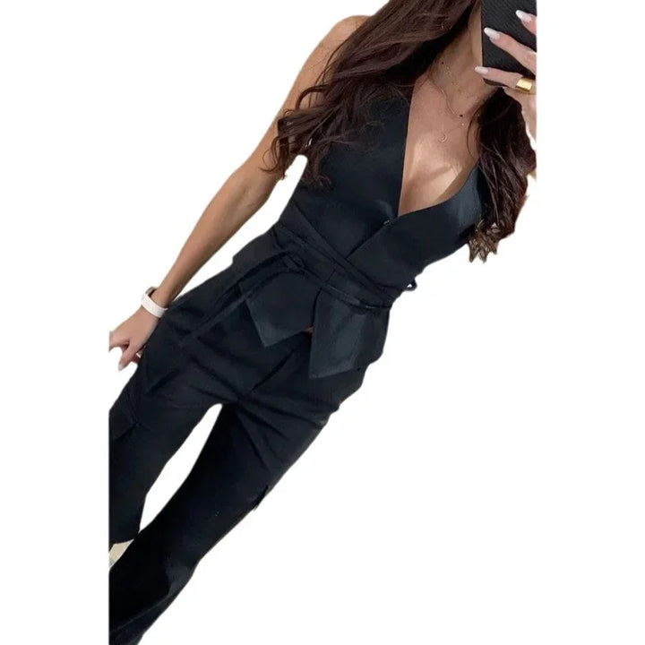 Woman wearing a black sleeveless jumpsuit with deep V-neck and belt detail, posing with phone