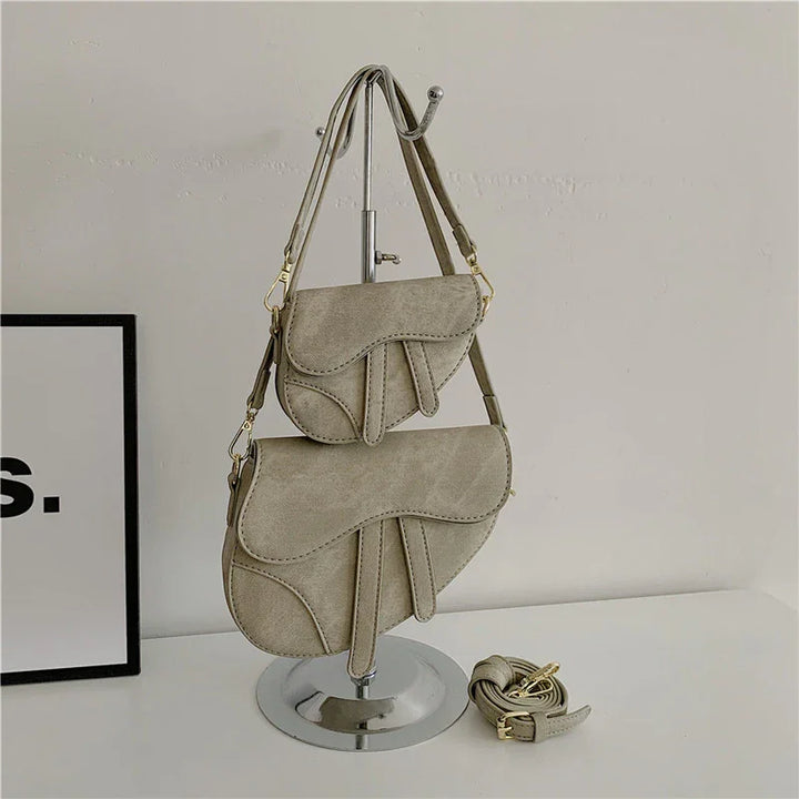 Beige suede saddle handbags displayed on metal stand with detachable strap on white surface