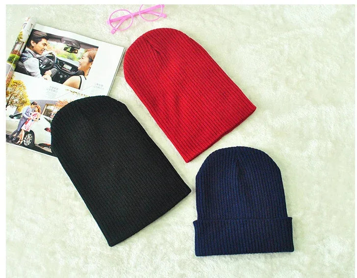 Three ribbed knit beanies in black, red, and navy blue on a white fuzzy surface with an open magazine and pink glasses