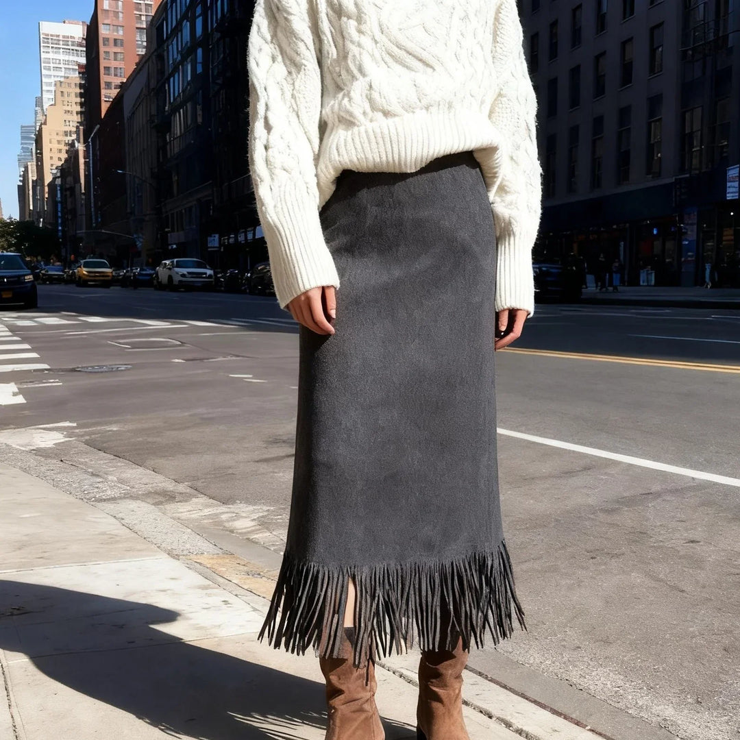 Woman wearing a cream cable knit sweater and gray suede fringe midi skirt with brown suede boots on city street
