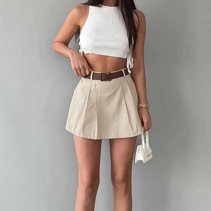 Woman wearing white ribbed crop top and beige high-waisted mini skirt with brown belt holding small white handbag