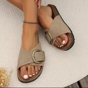 Close-up of feet wearing beige slip-on sandals with large buckle on wooden deck