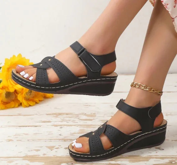 Black wedge sandals with ankle straps worn on feet against white background with sunflowers