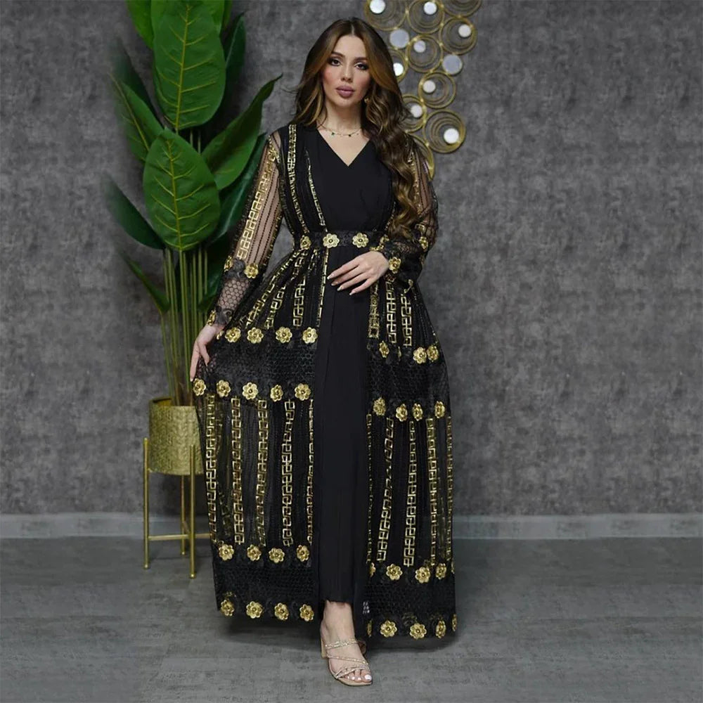 Woman in black and gold embroidered long dress posing indoors with green plant and decorative wall art