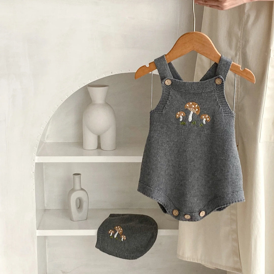 Gray knitted baby romper with mushroom embroidery and matching hat on white shelves
