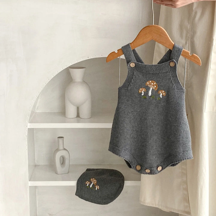 Gray knitted baby romper with mushroom embroidery and matching hat on white shelves