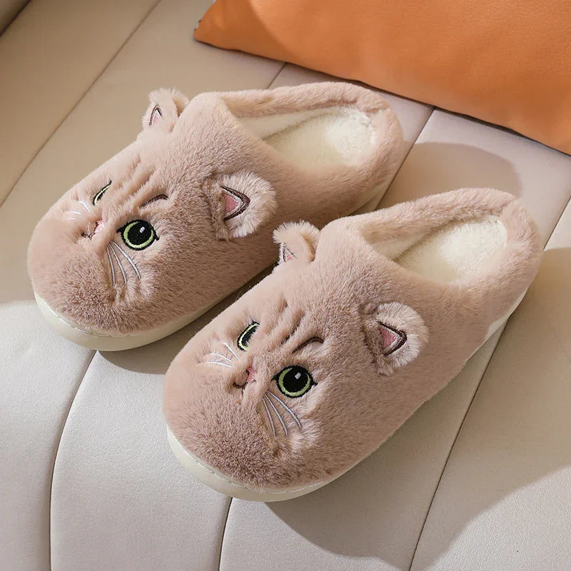 Cozy beige fuzzy slippers with embroidered cat face and ears, placed on cream sofa with orange pillow