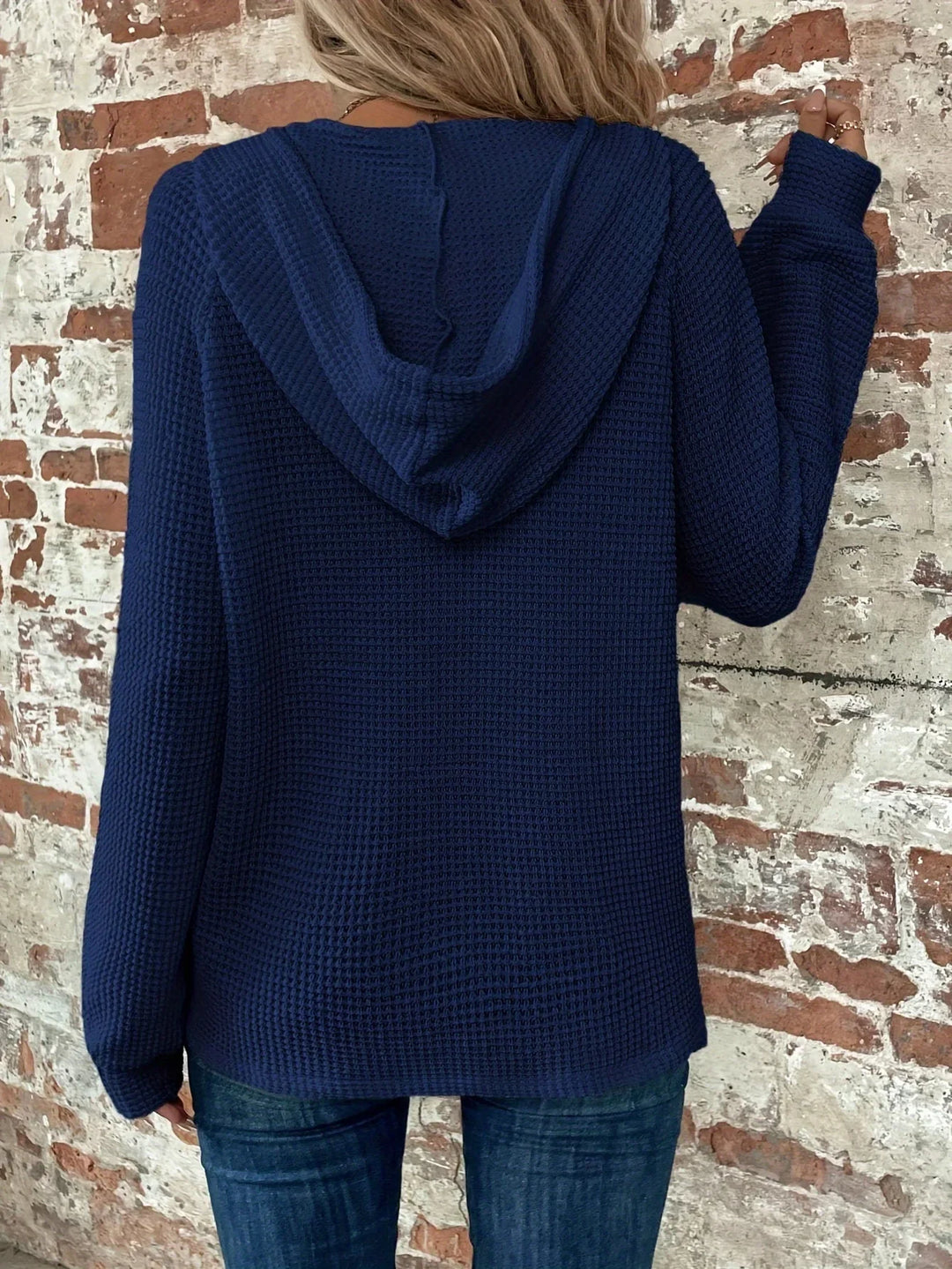 Woman wearing navy blue waffle knit hoodie with hood up, standing against rustic brick wall