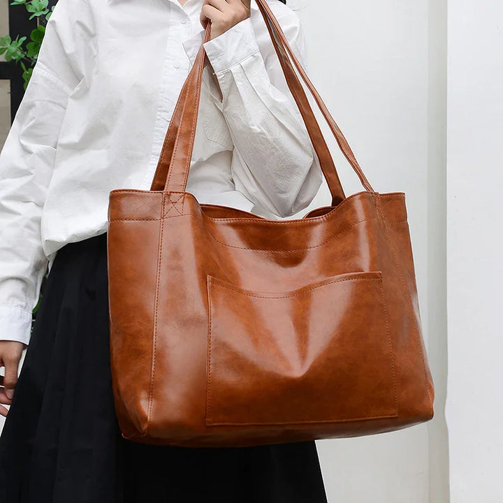Woman holding large brown leather tote bag with front pocket, wearing white shirt and black skirt