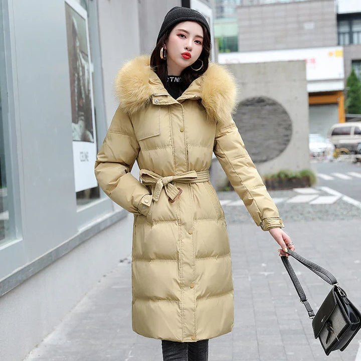 Woman wearing beige belted winter coat with fur hood and black beanie, holding black handbag outdoors