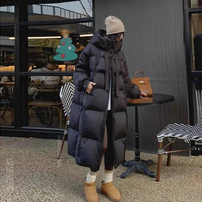 Woman wearing long black puffer coat, beige beanie, and boots standing by outdoor cafe table with brown handbag