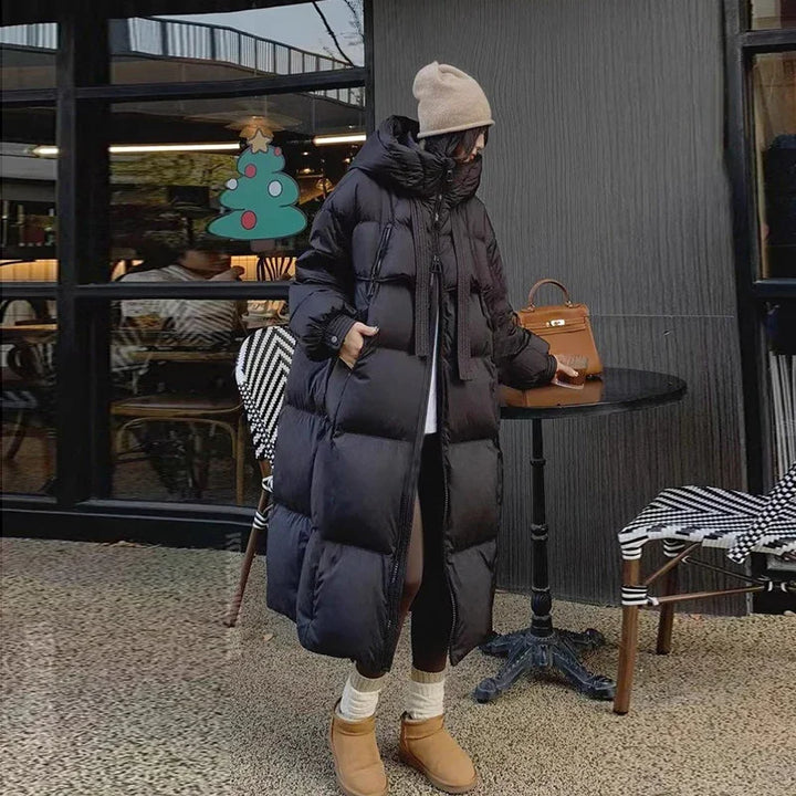 Woman wearing long black puffer coat, beige beanie, and boots standing by outdoor cafe table with brown handbag