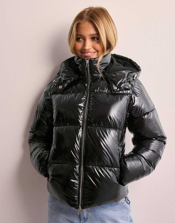 Smiling woman wearing shiny black puffer jacket with hood and light blue jeans against plain wall