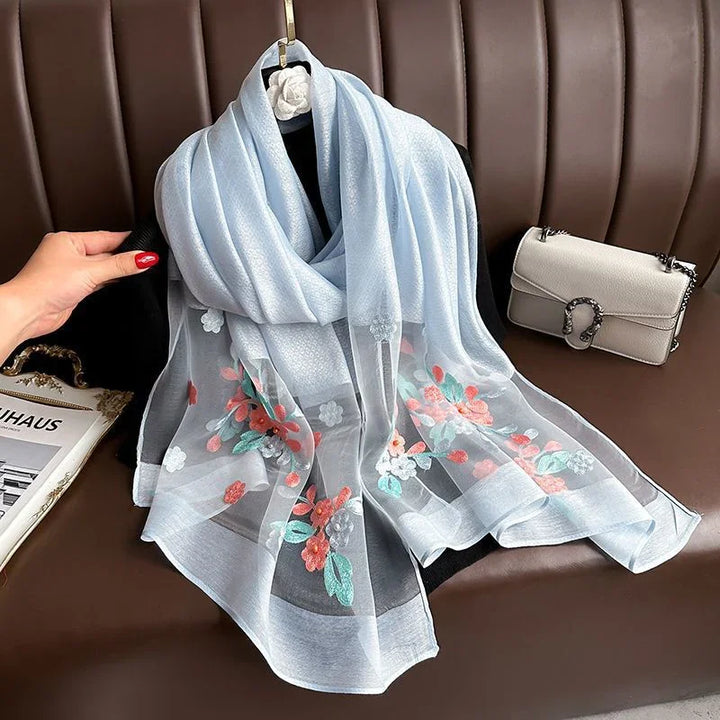 Light blue floral embroidered scarf displayed on black hanger over brown leather couch with beige handbag and magazine nearby