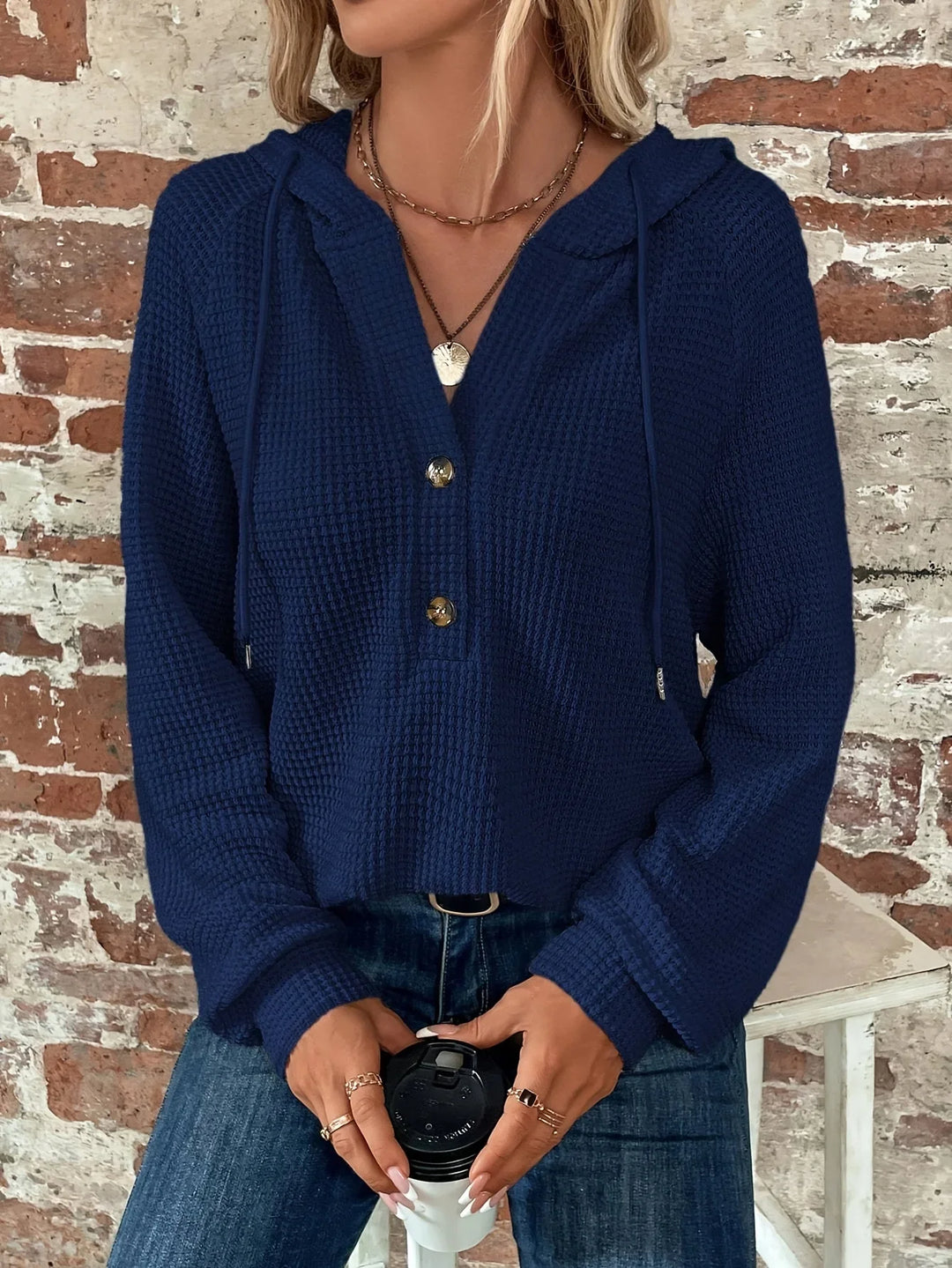 Woman wearing navy blue textured hooded sweater with buttons, holding coffee cup, brick wall background