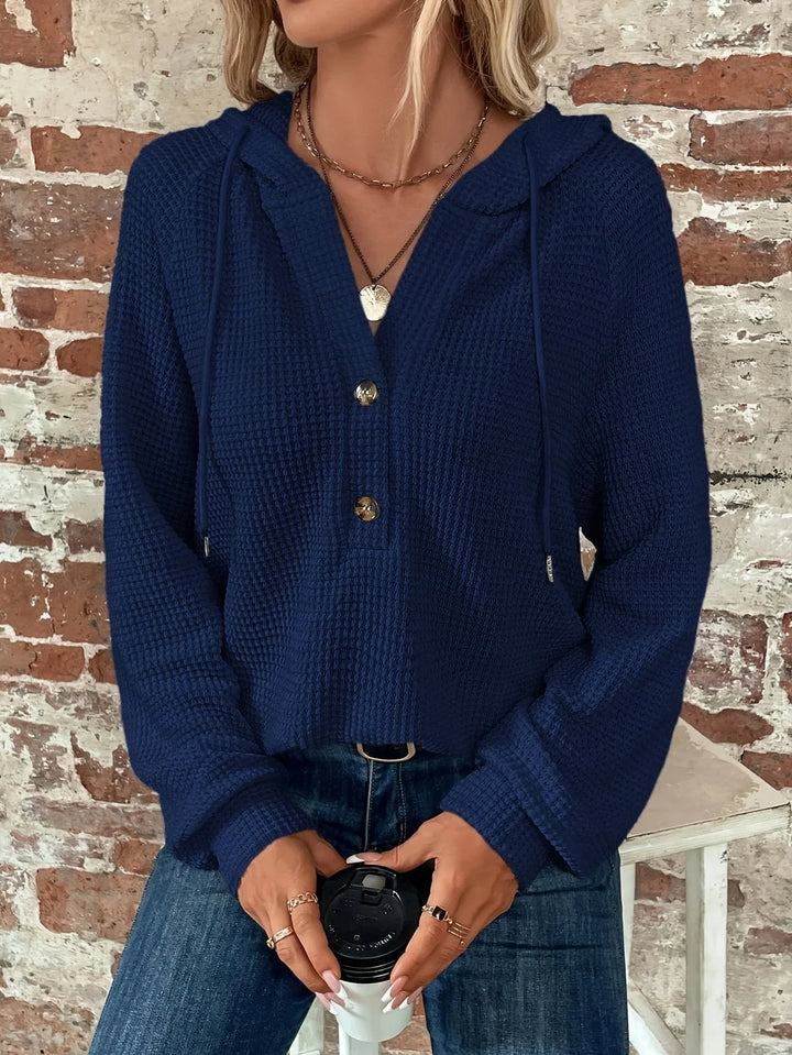 Woman wearing navy blue textured hooded sweater with buttons, holding coffee cup, brick wall background